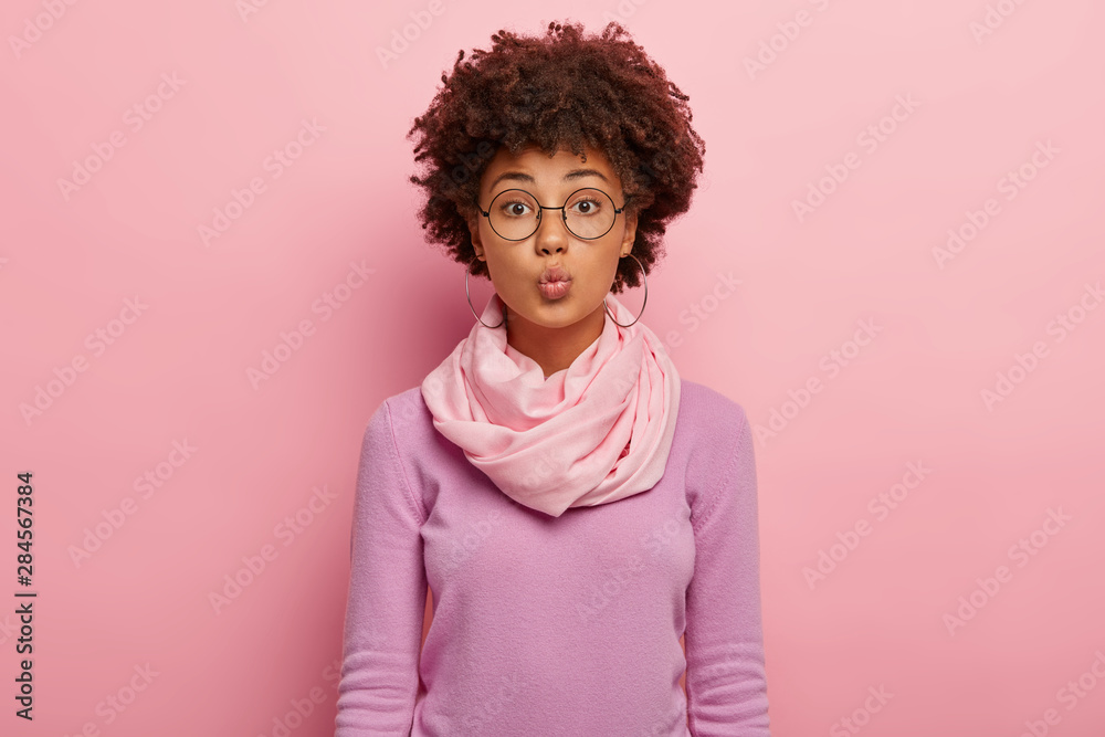 Tender feminine woman keeps lips folded, wants to kiss boyfriend, wears spectacles, hoop earrings, silk scarf around neck, looks straightly at camera, isolated on rosy wall. Human facial expressions Tender feminine woman keeps lips folded, wants to kiss boyfriend, wears spectacles, hoop earrings, silk scarf around neck, looks straightly at camera, isolated on rosy wall. Human facial expressions