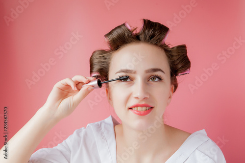 nice cheerful young the girl in the curler paints an eyebrow on pink background