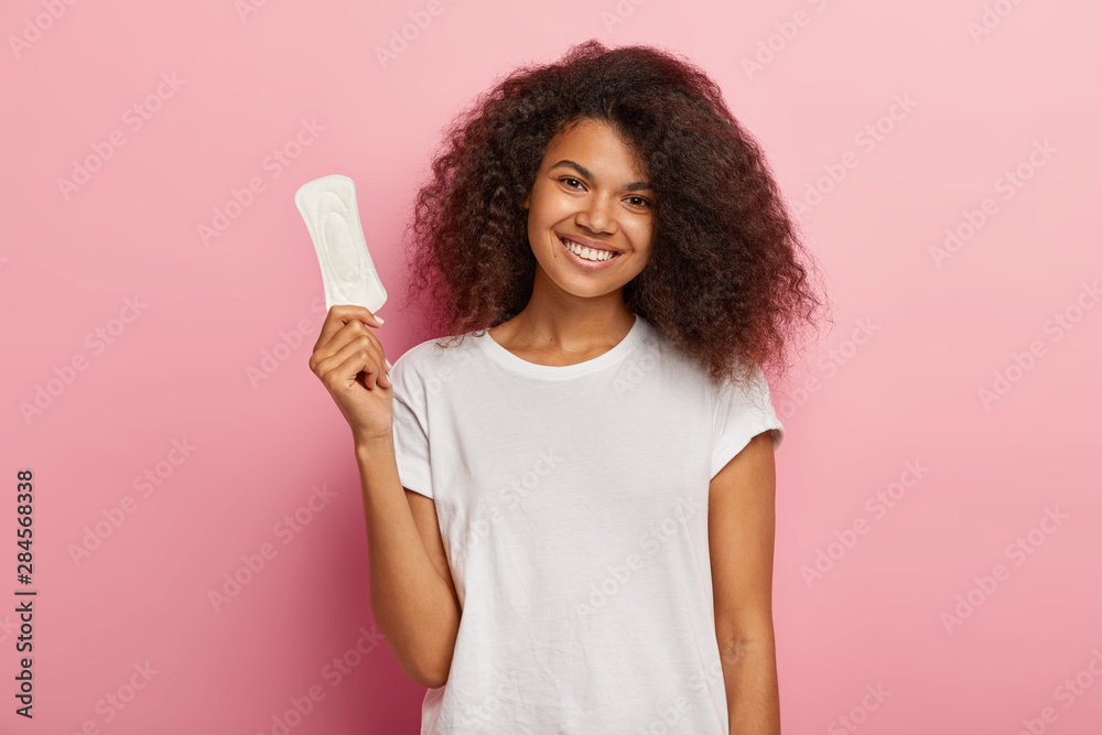 Glad attractive curly female model holds thin sanitary pad, uses feminine intimate product during menstruation or periods, cares about hygiene, wears casual t shirt, models over pink background - obrazy, fototapety, plakaty 
