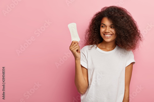 Satisfied lovely woman with crisp hairstyle, holds sanitary napkin, uses hygienic product during critical days, smiles positively, wears mockup t shirt, isolated on pink wall. Women health concept photo