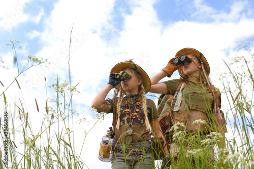 Two young girls seem to be on safari.They observe  the outdoor countryside area. They are dressed with  safari hats and khaki safari clothes.