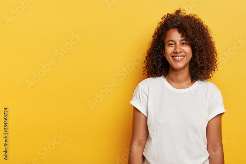 Relaxed female model with curly Afro hairstyle, grins from happiness, glad to have successful day, looks straightly at camera, wears white t shirt, laughs indoor over yellow background, copy space photo