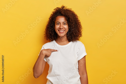 Smiling cheerful dark skinned girl points at herself, shows mockup space on white t shirt, happy being picked, models against yellow background. Carefree delighted young Afro woman asks who me