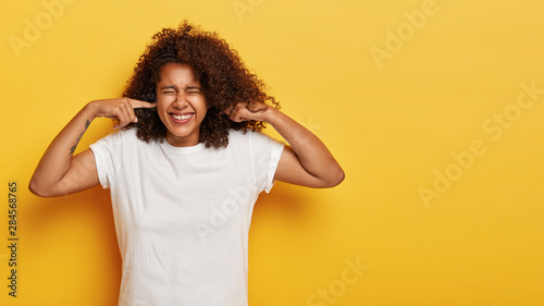 Studio shot of lovely stressed African American woman clenches teeth, doesnt want to listen anything, hears heartbreaking scream, plugs ears with index fingers, wears white t shirt, isolated on yellow