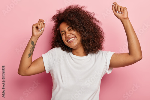 Funny joyous woman raises arms and dances carefree, feels pleasure and amused, laughs happily, eyes closed from satisfaction, moves along with music, has tattoo dressed in casual wear isolated on pink photo