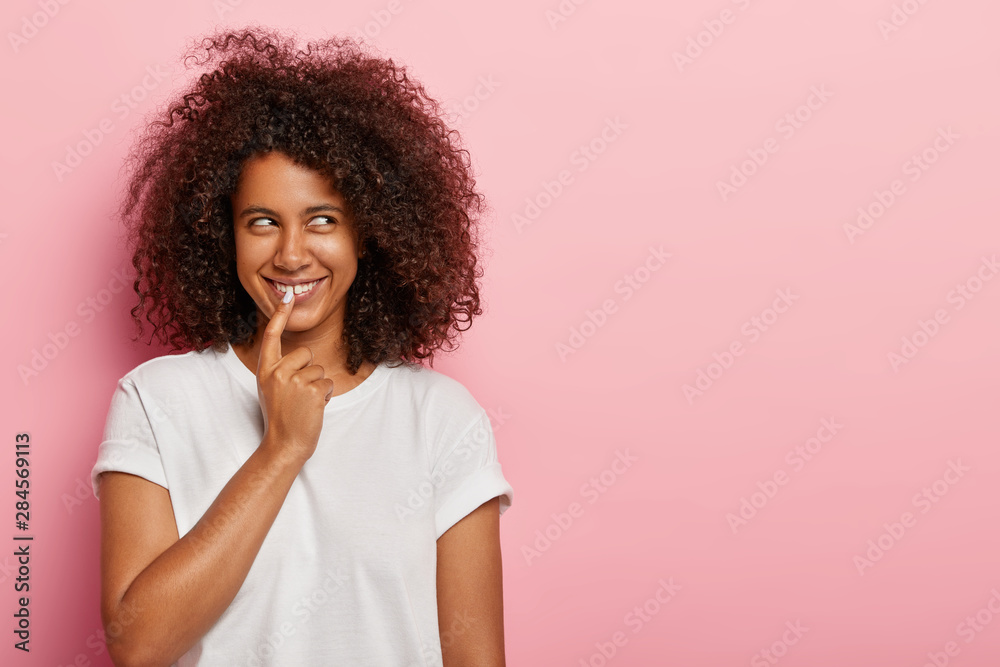 Studio shot of lovely delighted girl has intriuguing plan or intention to do something, keeps index finger on lips, has healthy dark skin, curly hairstyle, isolated on pink wall, blank space area Studio shot of lovely delighted girl has intriuguing plan or intention to do something, keeps index finger on lips, has healthy dark skin, curly hairstyle, isolated on pink wall, blank space area
