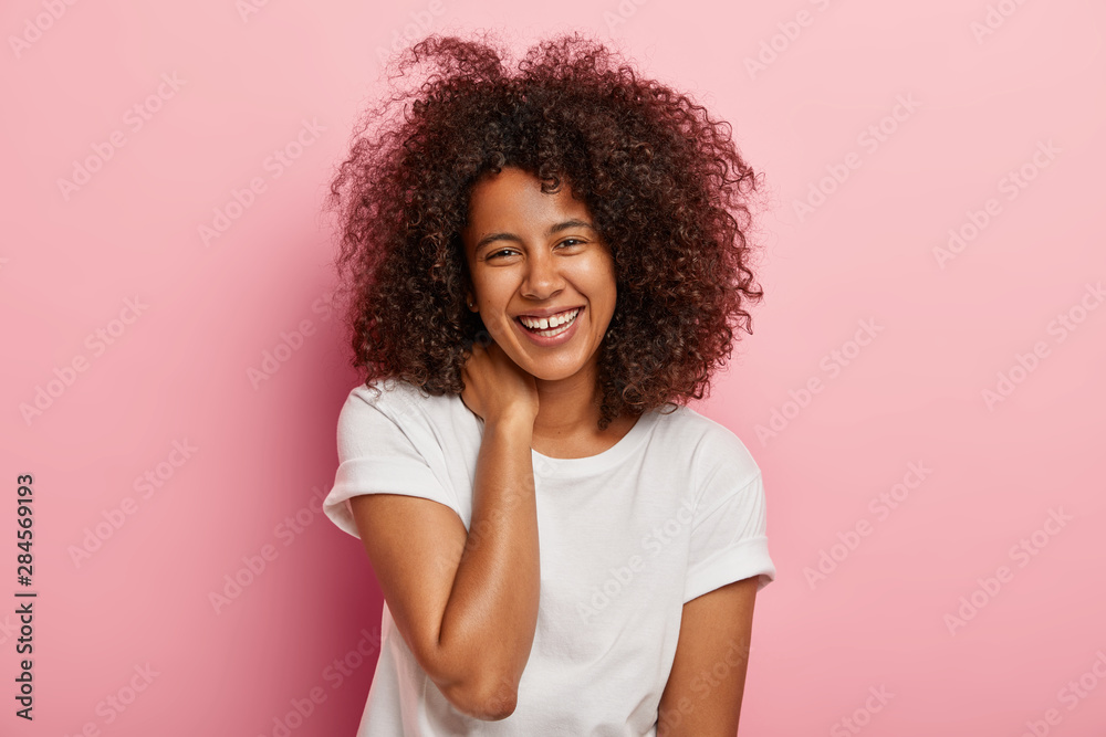 Glad dark skinned female has funny conversation, looks with broad smile at camera, entertained by friend, has overjoyed face expression, dressed in everyday clothes, stands indoor. Emotions concept Glad dark skinned female has funny conversation, looks with broad smile at camera, entertained by friend, has overjoyed face expression, dressed in everyday clothes, stands indoor. Emotions concept