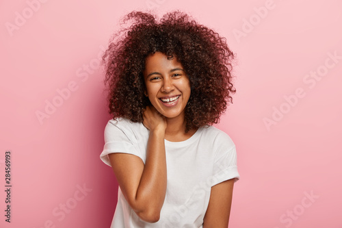 Glad dark skinned female has funny conversation, looks with broad smile at camera, entertained by friend, has overjoyed face expression, dressed in everyday clothes, stands indoor. Emotions concept