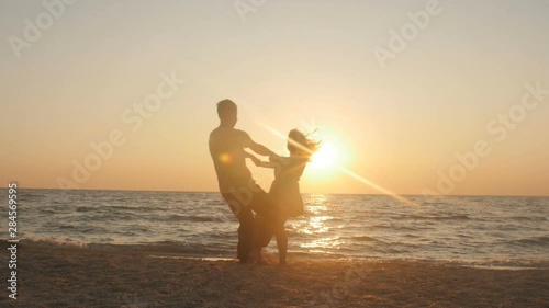 happy young couple on the beach, man spinning around his woman on sunset holding her arms at beautiful seaside. Having fun together at vacation. Slow motion