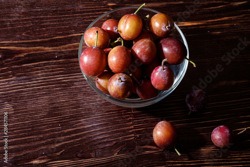 Ripe sweet plum fruits in glass bowl near with scattered plums on dark moody wood table background, hard light, copy space