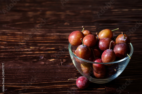 Ripe sweet plum fruits in glass bowl near with scattered plums on dark moody wood table background, hard light, copy space