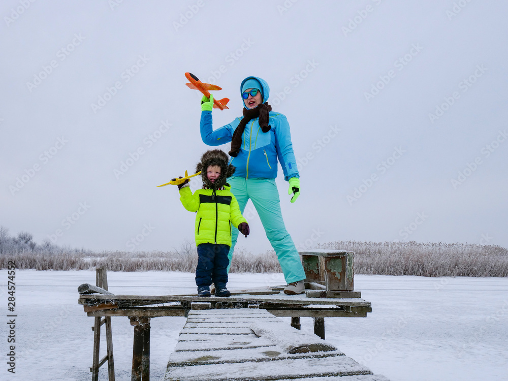 Slender cheerful mother in blue ski suit plays with children on snowy river. Boys with mom launch bright toy planes. Christmas Eve. Winter holidays in village. Family holidays.