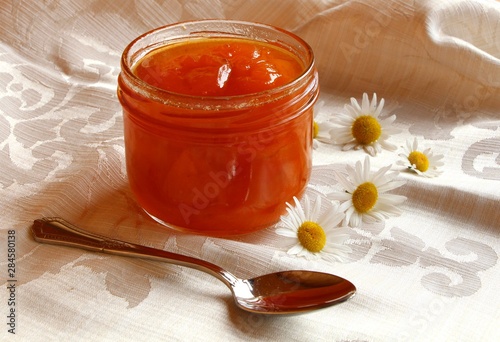 Closeup of  homemade  organic apricot jam in  an  open glass jar  on the background of a white tablecloth with daisy flowers and tea spoon.