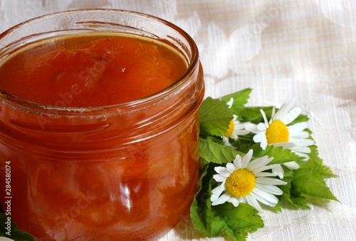 Closeup of  homemade  organic apricot jam in  an  open glass jar  on the background of a white tablecloth with daisy flowers. 