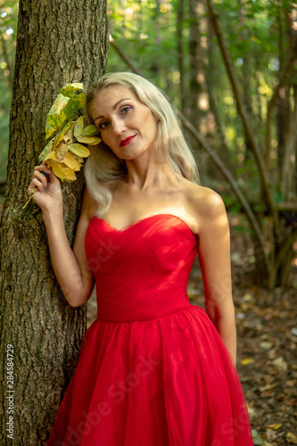 Fairy tale forest. A beautiful girl is standing in a long red dress near a tree and in her hand is holding a maple yellow leaf. Autumn forest.