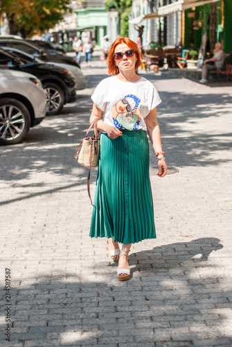 A young girl with red hair in a white T-shirt and a bright green skirt in sunglasses from the sun on her face is walking along the city street.