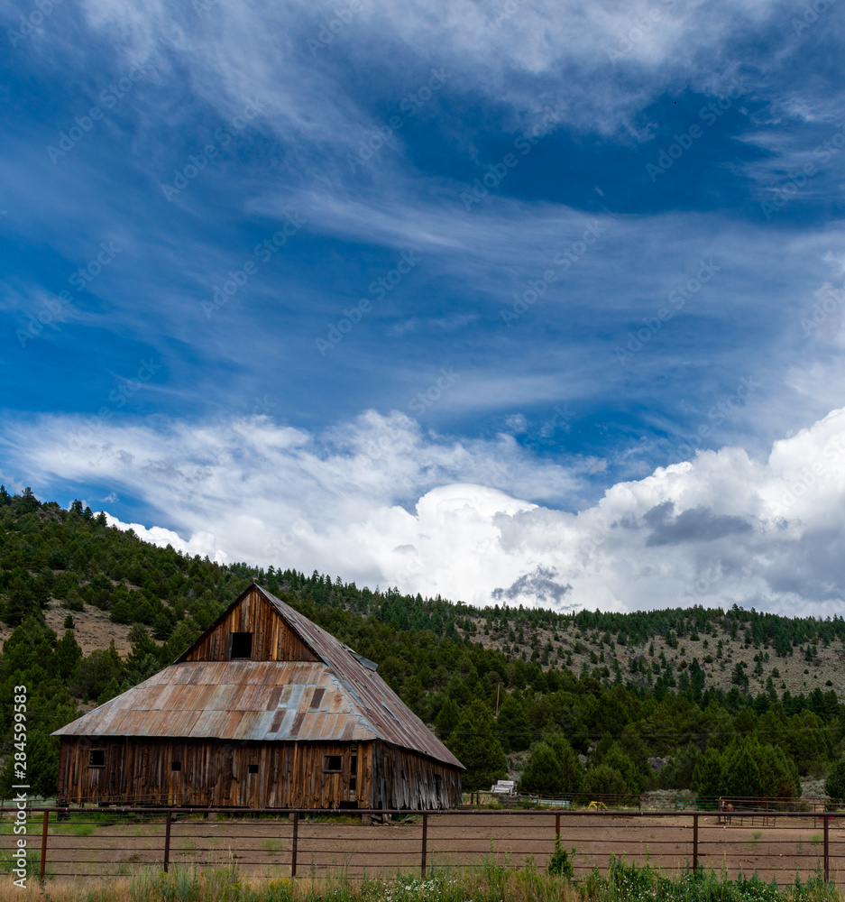 An ancient, weathered barn with rusted metal roof is backed by a forested hill under a vivid blue sky with billowing white clouds. An ancient, weathered barn with rusted metal roof is backed by a forested hill under a vivid blue sky with billowing white clouds.