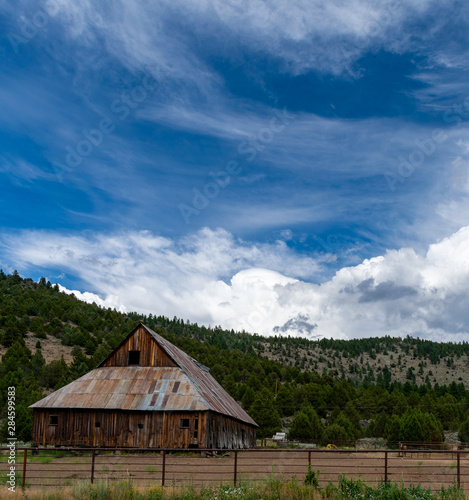 An ancient, weathered barn with rusted metal roof is backed by a forested hill under a vivid blue sky with billowing white clouds.