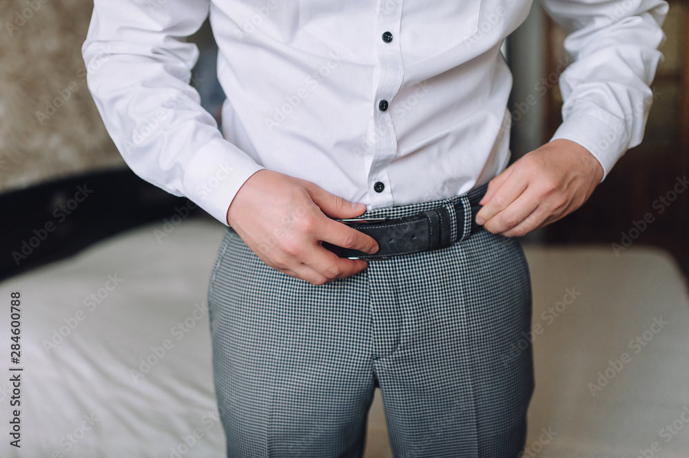 Fototapeta premium Groom in gray pants and a white shirt fastens his leather belt. Morning groom. Wedding details. Concept and composition. Businessman man and business.