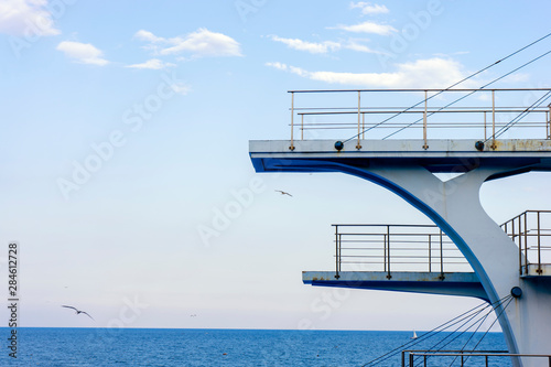 White diving board or tower against a clear blue sky. In the background the sea.