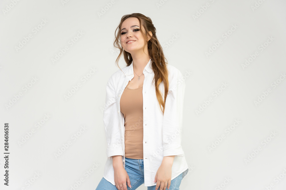 Photo portrait above the knee of a pretty brunette woman girl with long beautiful curly hair on a white background in a white shirt and blue jeans. Talking while standing in front of the camera. Photo portrait above the knee of a pretty brunette woman girl with long beautiful curly hair on a white background in a white shirt and blue jeans. Talking while standing in front of the camera.