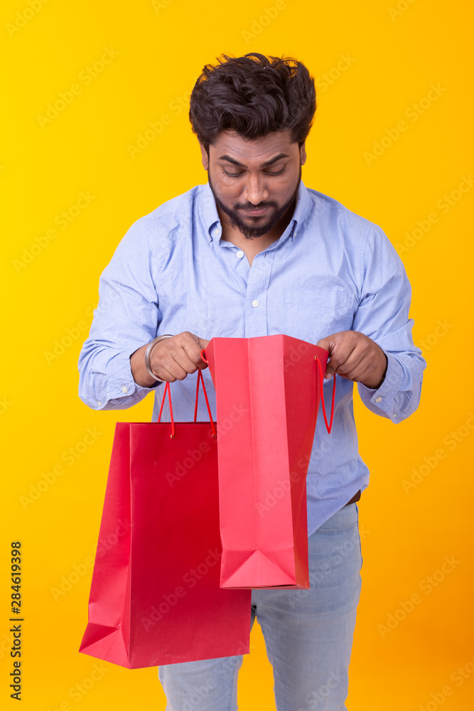 Surprised joyful young Indian man looks into the packages for shopping, standing on a yellow background. The concept of successful purchases.