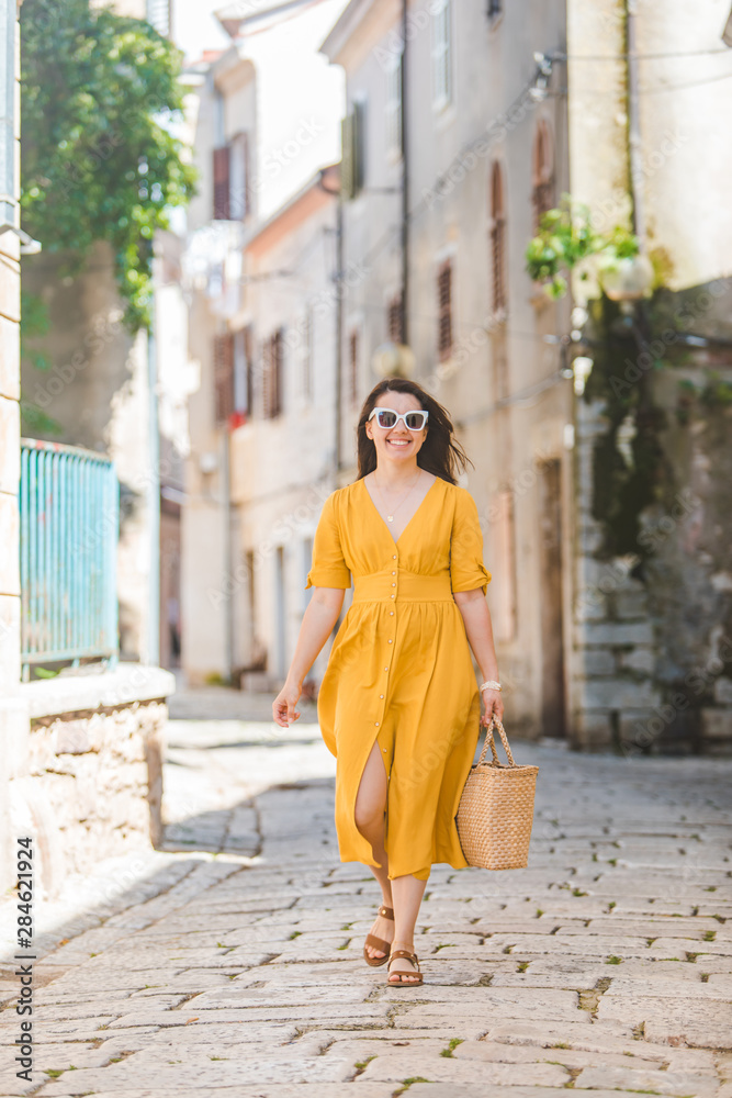 woman in yellow dress with bit straw hat walking by old tourist city in croatia