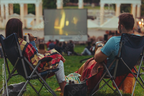 couple sitting in camp-chairs in city park looking movie outdoors at open air cinema