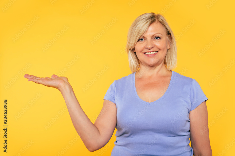 Fototapeta premium Happy excited beautiful blonde senior woman in casual clothes is pointing away on yellow background.