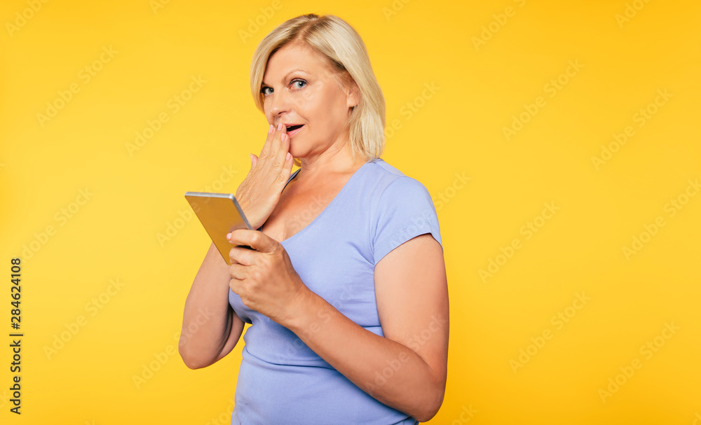Smiling beautiful cute blonde senior woman is using smart phone isolated on yellow background Smiling beautiful cute blonde senior woman is using smart phone isolated on yellow background