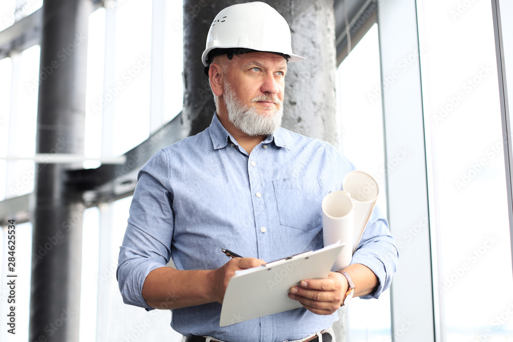 Confident mature business man in hardhat holding blueprint and looking away while standing indoors. - obrazy, fototapety, plakaty 