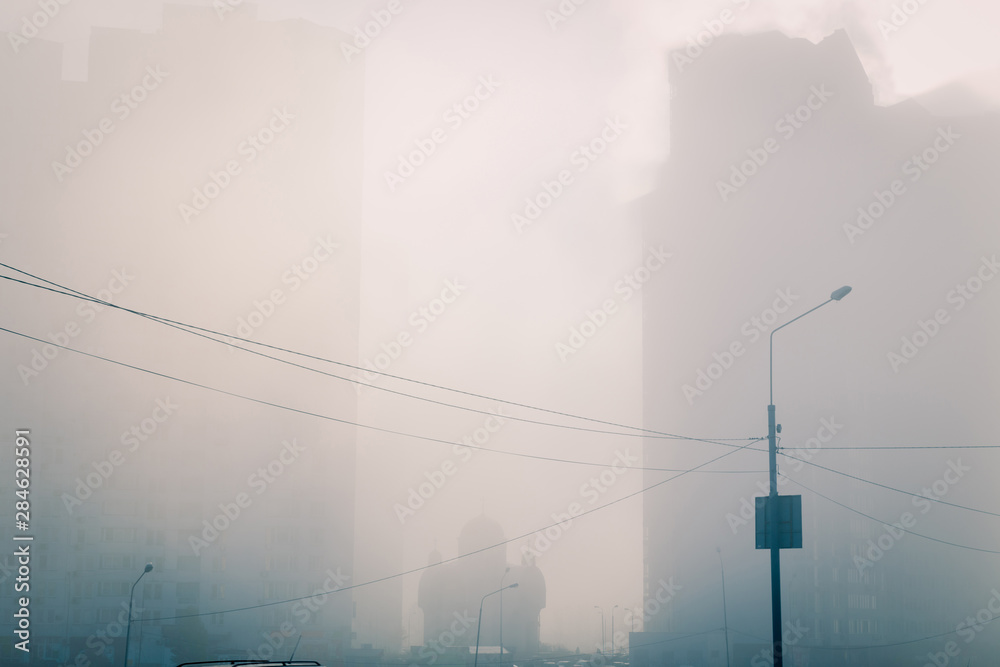 Dreamy city view. Small orthodox church silhouette in sun light, between high modern residential buildings, covered of dense morning fog. Kyiv. Ukraine.
