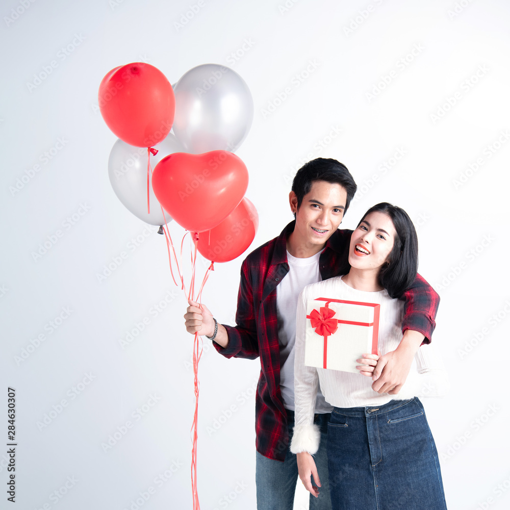 Beautiful young couple at home. Hugging, kissing and enjoying spending time together while celebrating Saint Valentine's Day with gift box in hand and air balloons in shape of heart on the background Beautiful young couple at home. Hugging, kissing and enjoying spending time together while celebrating Saint Valentine's Day with gift box in hand and air balloons in shape of heart on the background