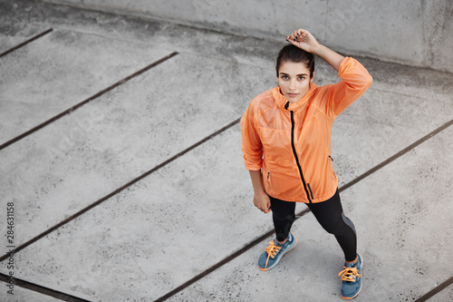 Upper view motivated confident young brunette woman in orange running jacket, looking up, standing concrete street outdoors, taking break as running, prepare marathon, jogging alone