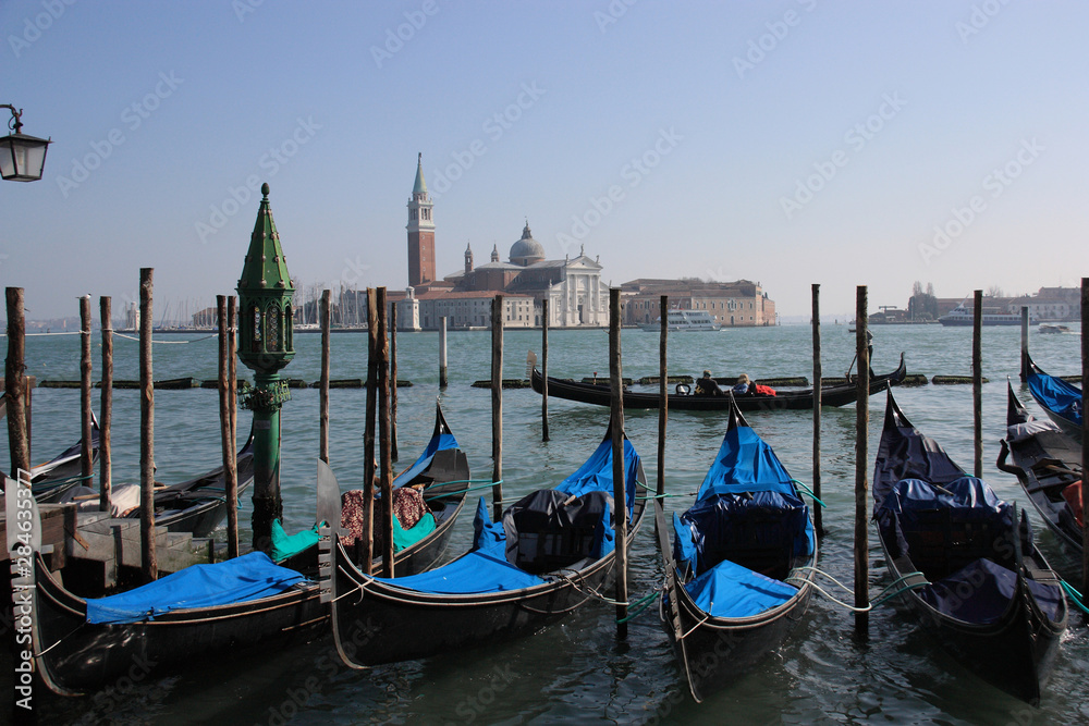 Venice, Italy. Rgand canal. Blue gondolas moored at a pier. Daylight, sunshine. Island in a distance. Flowing boat in a distance with gondolier and tourists (faces are hidden watching apart)