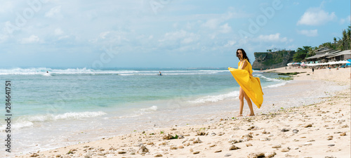 Woman in long yellow dress on rocky beach on Bali, Indonesia. Summer time. Dark hair brunette
