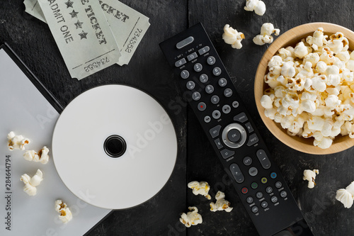DVD or blu ray movie disc with tv remote control, movie tickets and bowl of popcorn on dark background. Home theatre movie or series night concept. Flat lay from above.