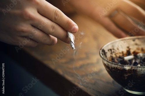 A man holds a Smoking cigarette in his hand, and a dirty glass ashtray stands on a wooden windowsill next to him.