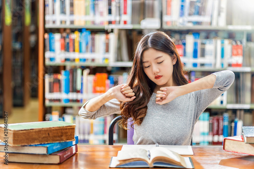 Asian young Student in casual suit reading and doing stretch oneself in library of university or colleage with various book and stationary on the wooden table over the book shelf, Back to school 