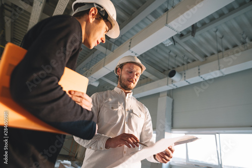 Architect and builder look on blueprint building under construction. Engineer and worker wearing safety hard hat have meeting on construction site