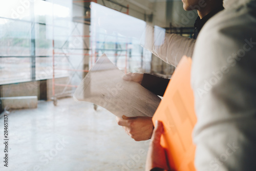 Architect and builder look on blueprint building under construction. Engineer and worker wearing safety hard hat have meeting on construction site