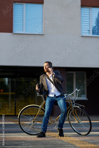 A young stylish bearded businessman in suit going to work by bike. Drinking coffee from a cup to go and talking on a mobile phone on the background of an office building. Takes a working break