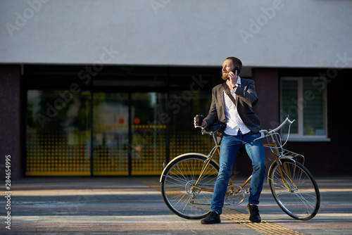 A young stylish bearded businessman in suit going to work by bike. Drinking coffee from a cup to go and talking on a mobile phone on the background of an office building. Takes a working break