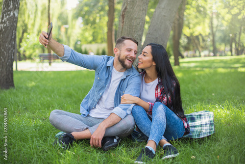 loving couple sitting on a plaid blanket in the park and use the phone, handsome guy and girl