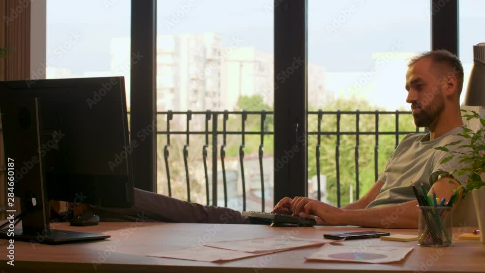 Businessman in start-up company office typing on PC with his feet on the desk. Warm sunset light coming from big windows Businessman in start-up company office typing on PC with his feet on the desk. Warm sunset light coming from big windows