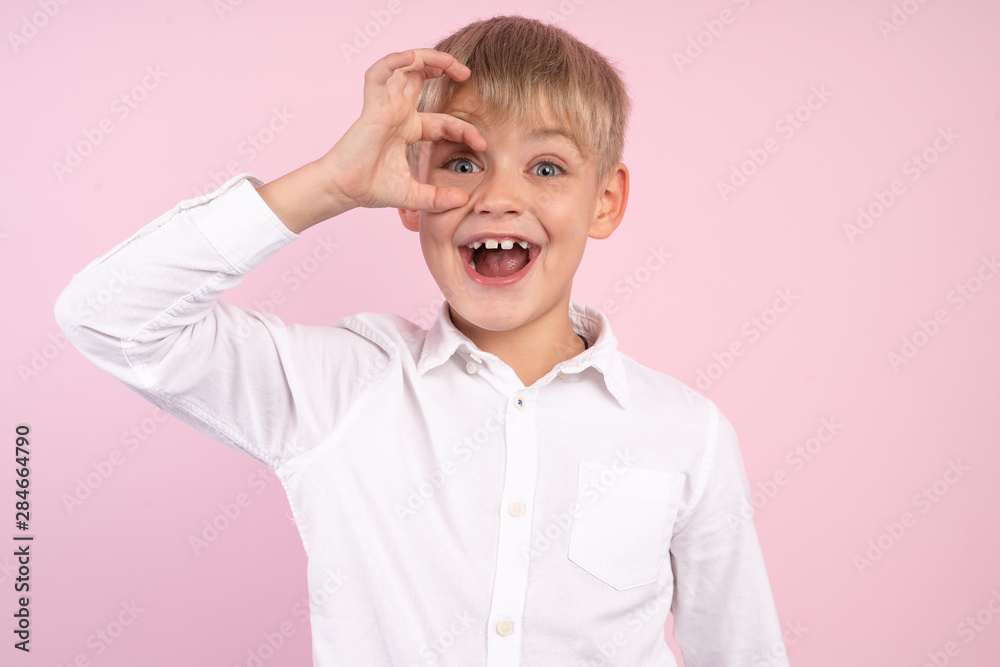 Portrait of little adorable cute boy having fun and smiling, holding fingers around his eyes. studio portrait over pink background. wearing white shirt. Portrait of little adorable cute boy having fun and smiling, holding fingers around his eyes. studio portrait over pink background. wearing white shirt.