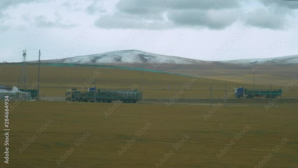 Two large cargo trucks speed down rural road crossing the plains of Tibet. Cinematic shot of large trucks carrying cargo across the vast Chinese countryside. Electricity wires obstructing the view. Two large cargo trucks speed down rural road crossing the plains of Tibet. Cinematic shot of large trucks carrying cargo across the vast Chinese countryside. Electricity wires obstructing the view.
