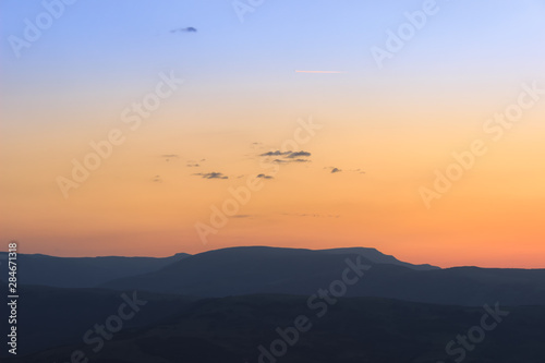 Cold to warm gradient on a sunrise sky above silhouette mountains, small dark clouds and sunlit plane trails