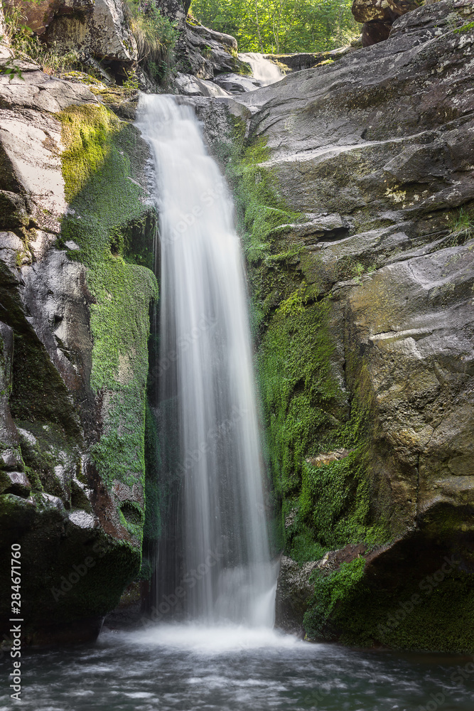 Obraz premium Scenic mountain waterfall cascading down the rocky cliff covered by colorful, vivid green moss and patches of sun hitting the rocks