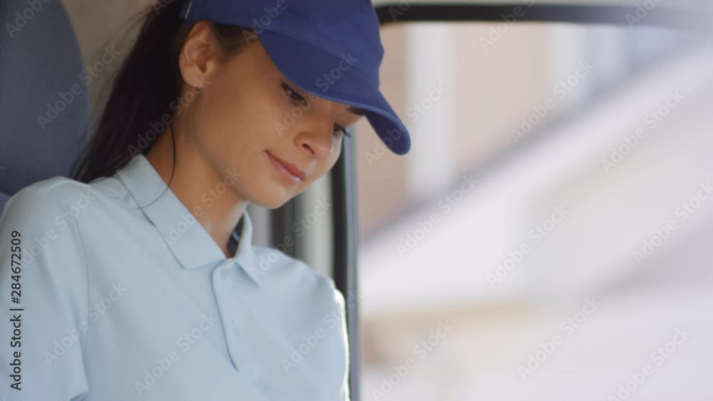 Handheld tilt up shot of cheerful young female courier sitting in drivers seat in delivery van and working on tablet